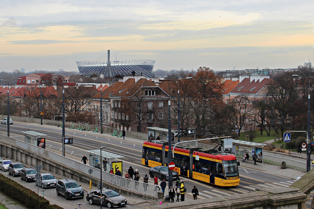Eine gelbe und schwarze Straßenbahn fährt auf einer von hohen Gebäuden gesäumten Stadtstraße, umgeben von Autos, Fußgängern, Laternen, Schildern, Bäumen und Gras unter einem bewölkten Himmel.