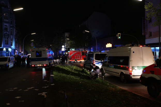 Eine Gruppe von Polizeiwagen steht nachts am Straßenrand, mit Menschen auf dem Bürgersteig, Gras, Pfählen, Laternen, Verkehrsampeln, Schildern, Bäumen, Gebäuden mit Fenstern und dem Himmel im Hintergrund.