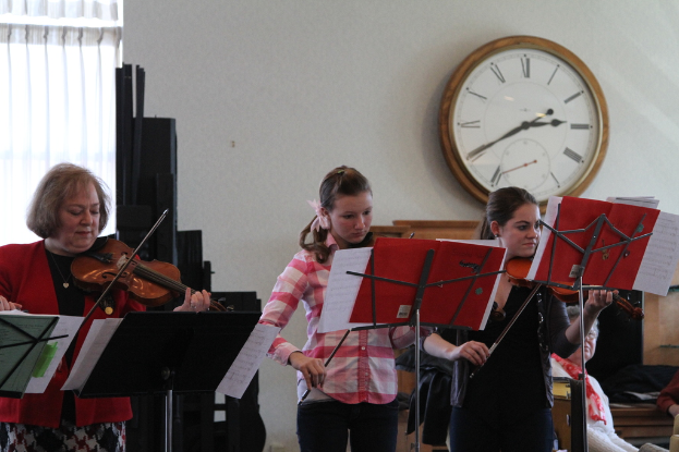 Drei Frauen, die Geigen spielen, vor Musikständern stehend, während eine Gruppe von sitzenden Frauen zuhört, vor einer weißen Wand mit einer Uhr und einem weißen Vorhang in der Ecke.