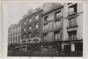 Schwarz-weiß-Foto einer Stadtstraße mit Gebäuden mit Fenstern, Geländern und Namensschildern sowie einer Straße unten.
