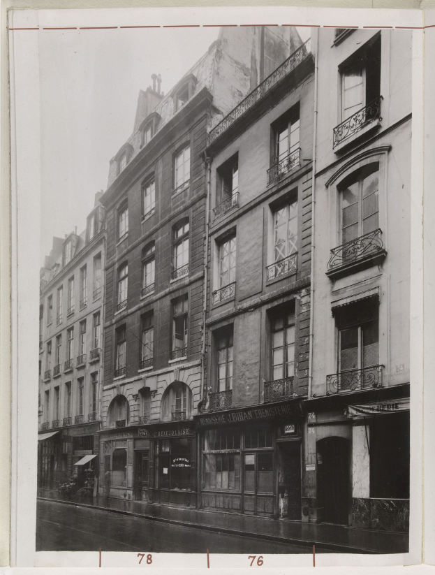 Schwarz-weiß-Foto einer Stadtstraße mit Gebäuden mit Fenstern, Geländern und Namensschildern sowie einer Straße unten.