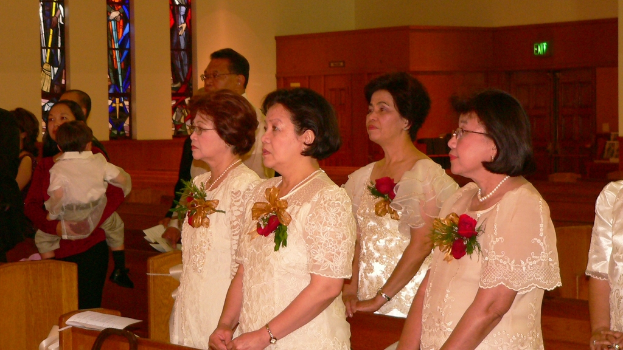 In der Mitte sind einige Frauen in Kleidern mit Blumen zu sehen, vor den Bänken auf der linken Seite stehen einige Personen, auf einer der Bänke liegt ein Stück Papier und an der Wand oben ist eine Tür zu sehen.