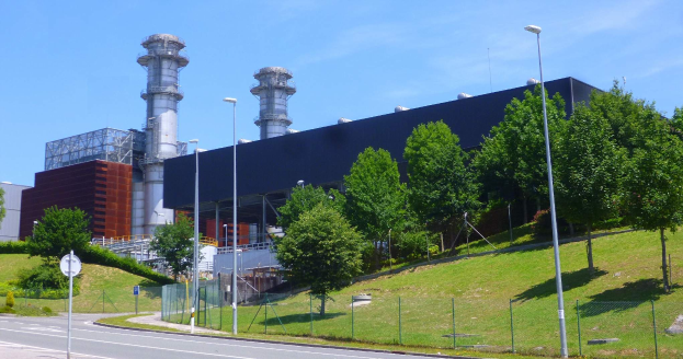 Ein großes Gebäude mit zahlreichen Fenstern, identifiziert als Kraftwerk durch Beschriftung, umgeben von Straßeninfrastruktur, Fahrzeugen, Grünfläche und einem bewölkten Himmel.