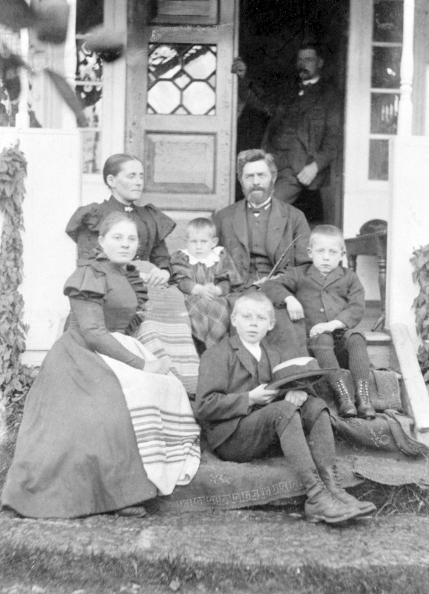 Ein Schwarz-Weiß-Foto einer Familie, die auf den Stufen eines Hauses sitzt, mit einem Mann im Hintergrund, das Haus hat eine Tür und Fenster.