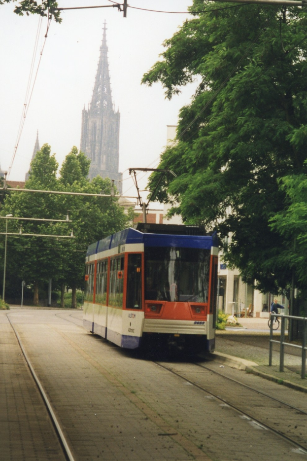 Ein roter und weißer Zug fährt auf Schienen neben einem hohen Gebäude, mit einem Radfahrer auf einem Fußweg rechts daneben; Bäume säumen die Schienen, und Gebäude und ein klarer blauer Himmel sind im Hintergrund zu sehen.