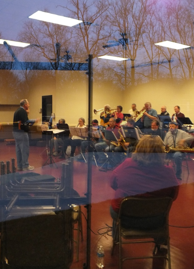 Musiker mit Trompeten im Hintergrund, Sänger vor Mikrofonen und ein Mann, der Musik komponiert, mit einer Wand und Bäumen im Glasreflex.