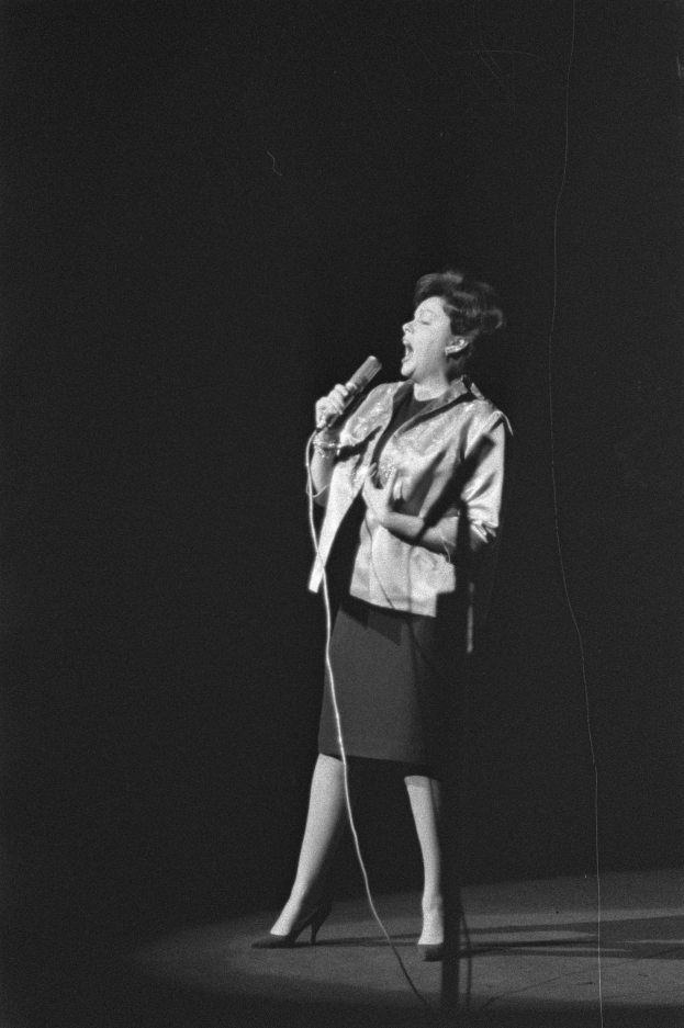 Eine Frau in einem schwarzen und weißen Outfit mit 1950er-Jahre-Frisur singt fröhlich auf einer von einem Spotlight beleuchteten Bühne und hält ein Mikrofon.