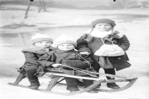 Drei Kinder in Jacken und Mützen sitzen auf einem hölzernen Schlitten im Schnee, mit Bäumen im Hintergrund.