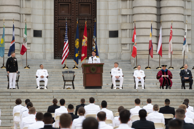 Eine Gruppe von Menschen in weißen Uniformen sitzt auf den Stufen eines Gebäudes, mit einem Podium und Flaggen davor und einem großen Gebäude mit einer Tür im Hintergrund.