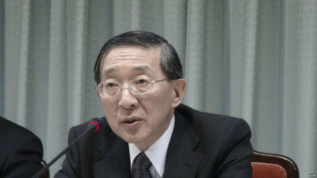 Ein Mann im Anzug und Krawatte sitzt an einem Tisch mit einem Mikrofon, trägt eine Brille und ist in einem formellen Setting mit einem Vorhang im Hintergrund zu sehen, während die Hand einer anderen Person links im Bild zu erkennen ist.