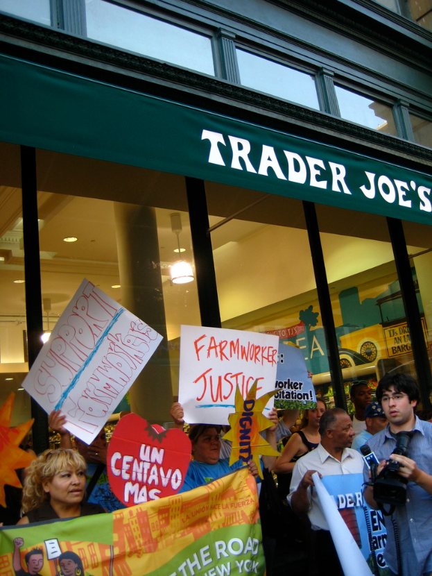 Eine Gruppe von Menschen steht vor einem Geschäft, hält Schilder und spielt Karten, mit einer Säule und Lampen auf dem Dach im Hintergrund
