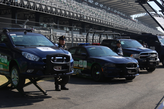 Polizeiautos vor einer Rennstrecke geparkt, uniformierte Personen auf der Straße, Stadion im Hintergrund und Maschendrahtzaun sichtbar.