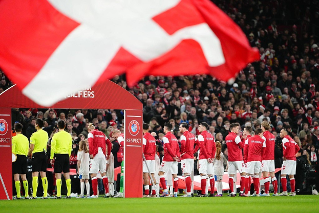 Gruppe von Menschen auf einem Fußballfeld mit einer roten und weißen Flagge im Vordergrund, einem Bogen mit der Aufschrift "Bayern München vs. Bayern München Betting Tips & Preview" im Hintergrund und einer großen Stadionmenge.