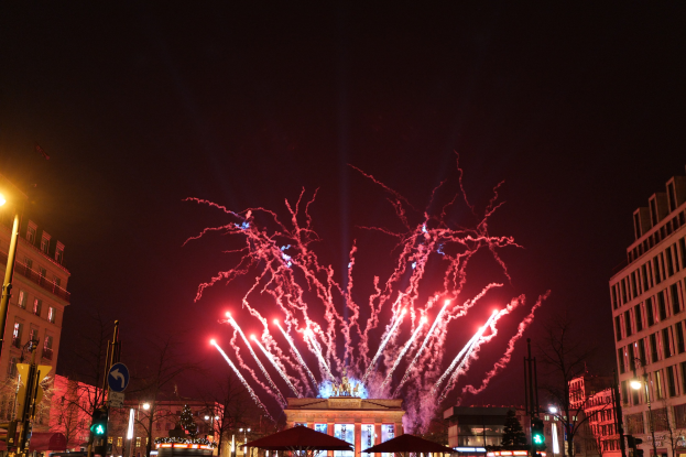 Eine belebte Stadtstraße am Neujahrstag in Berlin, voller Menschen, Fahrzeugen und Gebäuden, erleuchtet von Lichtern und Feuerwerken am Himmel.
