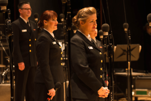 Eine Gruppe von Menschen, die vor Mikrofonen stehen, mit einem Mann und einer Frau, die Mikrofone halten, und Musikinstrumenten und Ständern im Hintergrund.