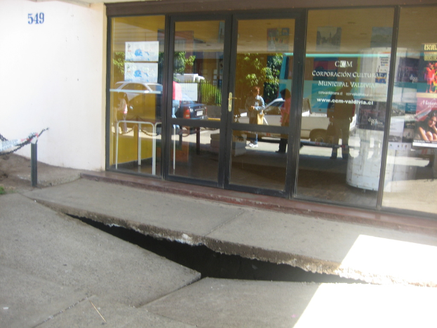 Eine gebrochene Oberfläche mit einer Glastür, Wand und einem Pfahl im Hintergrund, die sich bewegende Fahrzeuge und einige stehende Personen spiegeln.