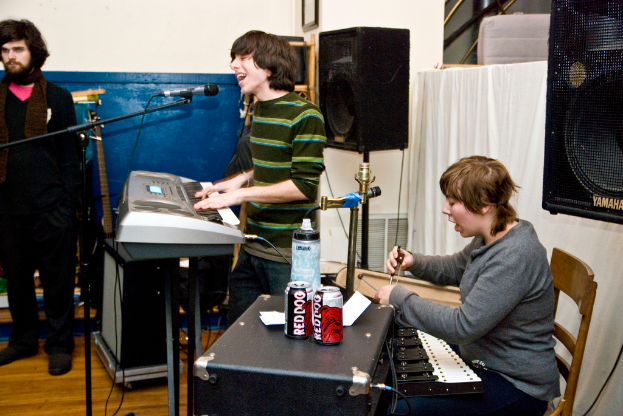 Drei Männer, einer steht links, einer spielt Klavier und singt, und einer singt, während er Dosen auf einem Tisch hält, mit einem Lautsprecher und einem Vorhang im Hintergrund.