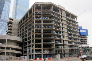 Großer Bau in einer städtischen Umgebung mit Wolkenkratzern, Fahrzeugen, Fußgängern, Verkehrsinfrastruktur, Kränen und einem klaren Himmel.