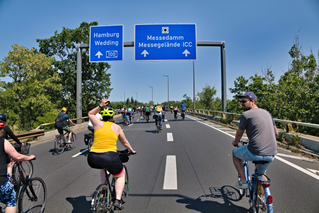 Gruppe von Radfahrern mit Helmen, die eine Straße mit Geländer und Bäumen auf beiden Seiten hinunterfahren, Laternen im Hintergrund und ein klarer blauer Himmel, mit einem Schild, das eine Radtour in Hamburg anzeigt.