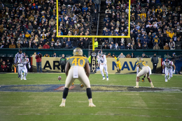 Fußballspieler mit Helmen und Sportbekleidung auf einem Feld mit Zuschauern auf den Rängen und Stadionwerbung im Hintergrund.