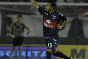 Ein Fußballspieler liegt auf dem Boden und feiert ein Tor, mit einer stehenden Person und einer Texttafel im Hintergrund und einer unscharfen Menge dahinter.