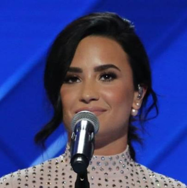 Demi Lovato steht selbstbewusst vor einem Mikrofon bei den Grammys, trägt ein schwarzes Kleid und eine stylische Bob-Frisur, mit einem blauen Hintergrund.