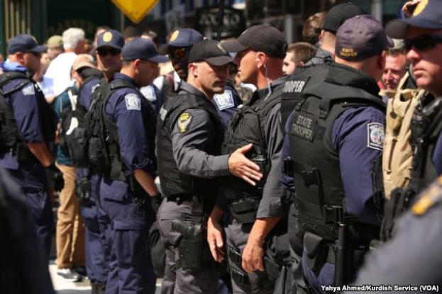 Eine Gruppe von Polizisten in Uniform steht in einer Reihe auf einer Straße, einige halten Waffen, mit Gebäuden und Schildern im Hintergrund.