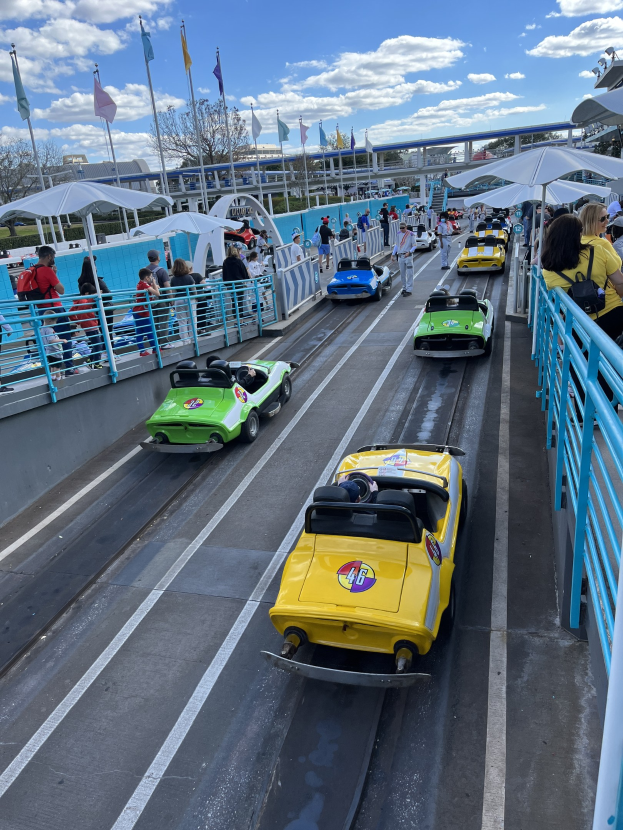 Gruppe von Menschen, die auf einer Achterbahn in Disney's Hollywood Studios fahren, umgeben von Geländern und Fahnen, mit Bäumen und Gebäuden im Hintergrund unter einem bewölkten Himmel.