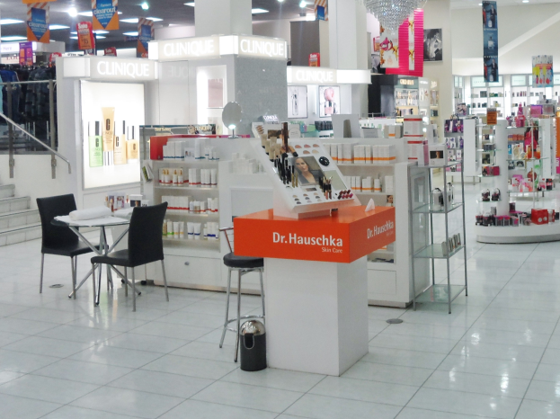 Ein gut beleuchteter Laden mit einer Vielzahl an Kosmetika, organisiertem Zubehör, Möbeln und Dekorationen, darunter Stühle, ein Tisch, ein Müllbehälter, eine Lampe, Schilder, eine Treppe, Kleidung und Deckenleuchten.