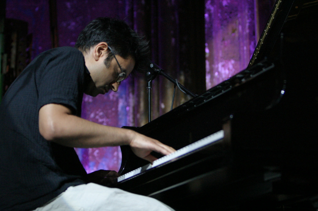 Ein Mann mit Brille spielt am Piano mit einem Mikrofonständer vor ihm und einer Wand im Hintergrund.
