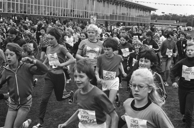 Schwarzes Foto von Kindern, die auf einem Rasenfeld mit einem Gebäude, Bäumen und Pfosten im Hintergrund unter einem klaren Himmel rennen.