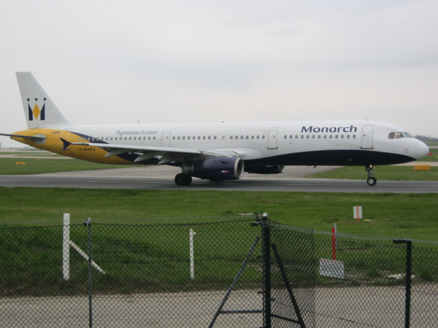 Eine Monarch Airlines Airbus A320-200 auf einer Landebahn, umgeben von Gras und einem Zaun mit Pfählen und Brettern, mit Bäumen und einem klaren blauen Himmel im Hintergrund.