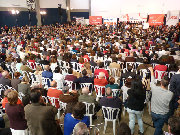 Ein volles Stadion mit sitzenden und stehenden Menschen, eine Bühne mit Individuals nahe einem Podium, Banner und Lichter.