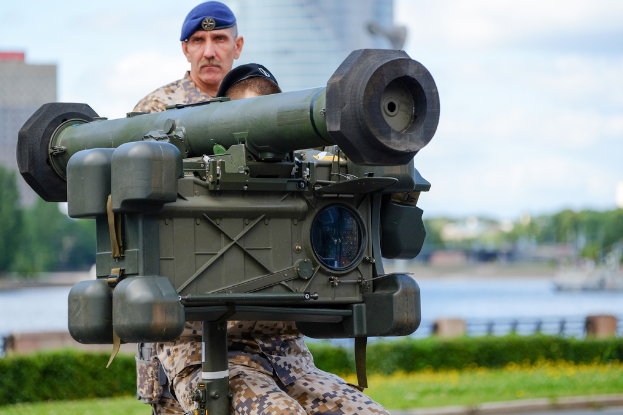 Zwei Personen in Militäruniformen mit grünen Waffen vor einem See mit Gebäuden, Bäumen und einem klaren Himmel
