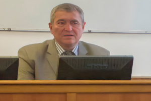 Ein Mann in einem Anzug sitzt an einem Schreibtisch vor einem Computer, mit einer Whiteboard im Hintergrund und sieht ernst aus und könnte ein Richter sein.