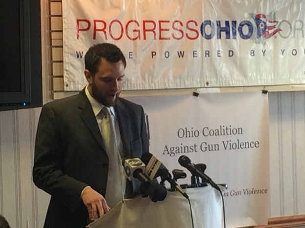 Eine Person steht auf einer Bühne vor einem Tisch mit Mikrofonen, mit einem Banner an der Wand dahinter.