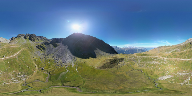 Panoramischer Blick auf eine Bergkette mit grünem Gras, einer gewundenen Straße und einem hellblauen Himmel unter einer strahlenden Sonne.