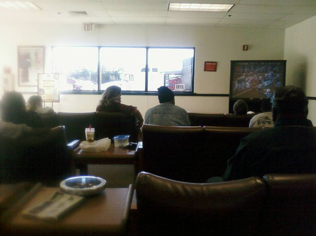 Ein Raum mit einer Decke, einem Fenster, einer Couch mit sitzenden Menschen, einem Tisch mit einer Tasse, einer Schachtel, einer Abdeckung, einem Buch und einer Schüssel, einem Banner an der Wand, einem Mann mit einer Mütze und einem Ausgangsschild an der Decke.