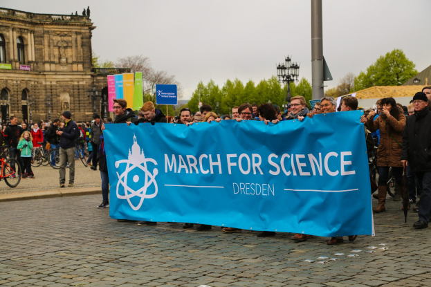 Eine Gruppe von Menschen hält eine 'March for Science Dresden'-Plakette vor einem Gebäude mit Bäumen, Fahrrädern und Laternen im Hintergrund.