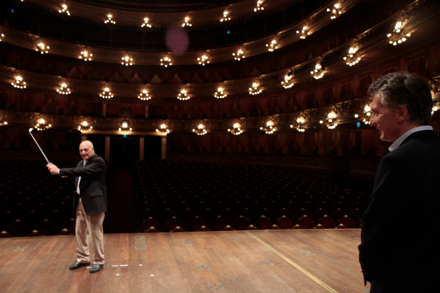Ein älterer Mann in einem Anzug hält einen Golfschläger auf der Bühne, neben ihm steht ein weiterer älterer Mann rechts, unter Deckenlampen mit vielen Sitzplätzen darunter.