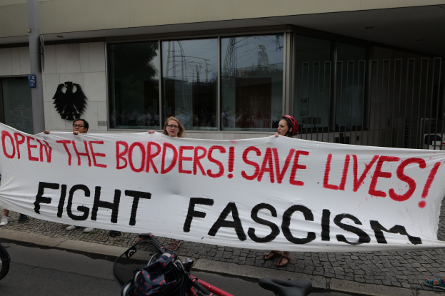 Demonstranten halten ein Transparent mit der Aufschrift "Öffnet die Grenzen, rettet Leben, kämpft gegen Faschismus" vor einem Gebäude mit Glasfenstern und einem Mast, mit einem Fahrrad und einer Tasche auf der Straße.
