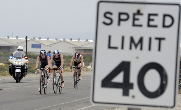 Eine Gruppe von Menschen in Helmen auf Fahrrädern auf einer Straße neben einem Geschwindigkeitsbegrenzungsschild, mit Gebäuden, Polen, Pflanzen, Bäumen und einem klaren blauen Himmel im Hintergrund.