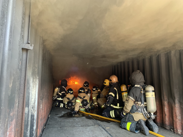 Feuerwehrleute mit Helmen und Sauerstoffgeräten arbeiten in einem raucherfüllten Tunnel, mit einem sichtbaren Feuer im Hintergrund und einer Wand auf der linken Seite.
