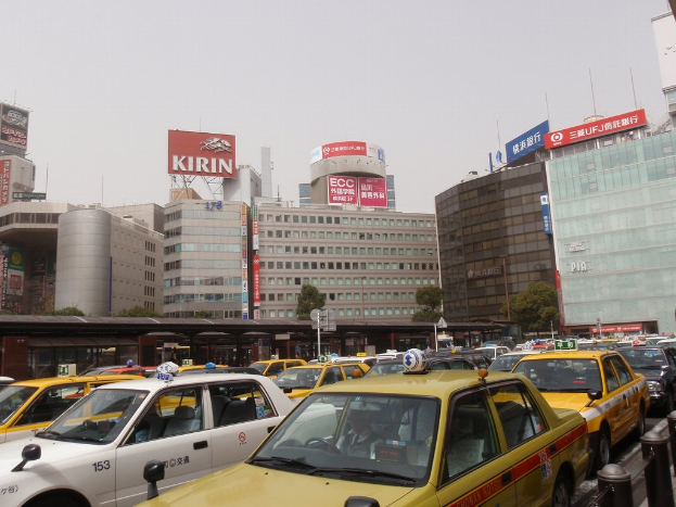 Eine Stadtansicht mit Fahrzeugen auf der Straße, Gebäuden, Bäumen, Polen mit Schildern und Text auf den Schildern, einem Zug im Hintergrund und einem Geländer unten rechts.