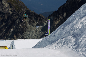 Ein Mann mit Skiern springt in der Luft mit schneebedeckten Bergen und einer Flagge mit Text im Hintergrund und einem stehenden Mann vor der Flagge, mit Text in der unteren linken Ecke.