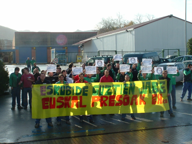 Eine große Gruppe von Menschen mit Papieren und einer Fahne im Vordergrund, mit Fahrzeugen, Geländern, Gebäuden, Bäumen und einem Schild im Hintergrund unter einem klaren Himmel.