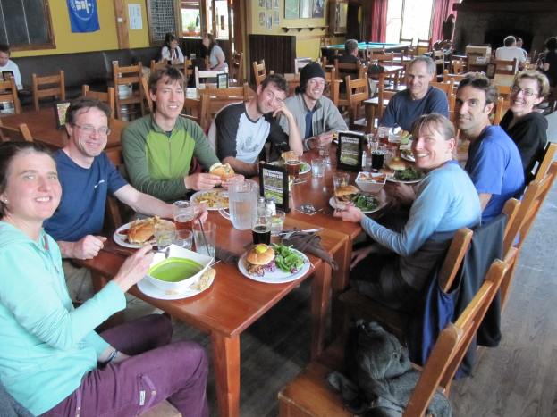 Eine Gruppe von Menschen sitzt um einen Tisch mit Tellern, Schalen, Gläsern, Flaschen, einem Wasserglas, Karten, Tuch und Essen, lachend, mit einer Jacke auf einem der Stühle, während einige weitere Personen an anderen Tischen im Hintergrund sitzen, mit einem Banner, Bildern und Postern an der Wand und einem Fenster mit Vorhängen.