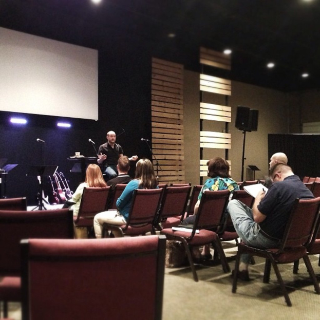 In einer Halle sitzen mehrere Frauen auf roten Ledersesseln, während ein Mann in einem schwarzen Hemd vor ihnen spricht. Hinter ihm ist ein weißer Projektionsschirm, hölzerne Wanddekorationen und ein Lautsprecher auf einem Ständer. Die Decke ist schwarz mit Scheinwerfern.