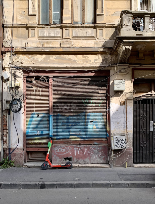 Ein roter E-Scooter neben einem Gebäude mit Fenstern, Rollläden und einem Tor auf einer Straße mit sichtbaren Kabeln und Gegenständen, einschließlich Text am unteren Rand.