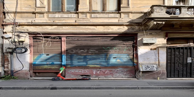 Ein roter E-Scooter neben einem Gebäude mit Fenstern, Rollläden und einem Tor auf einer Straße mit sichtbaren Kabeln und Gegenständen, einschließlich Text am unteren Rand.